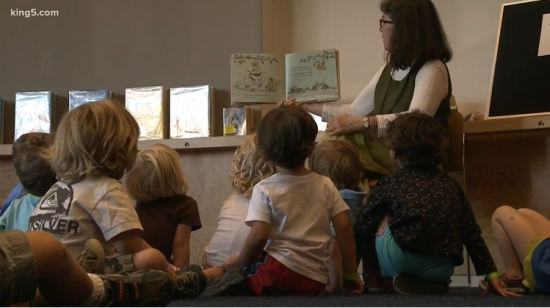 Children face away from the camera while they listen to a woman read aloud from a picture book.