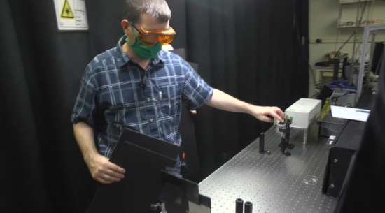 A man in a blue plaid shirt, green mask, and orange safety goggles holds a device over a metal table.