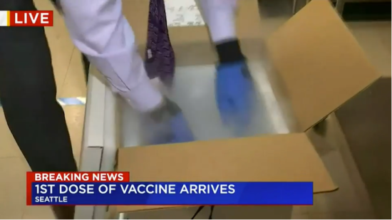 A brown cardborad box sits open on the floor. A person wearing blue latex gloves leans over and reaches into the box. A banner at the bottom of the screen reads: Breaking news. First does of vaccine arrives.