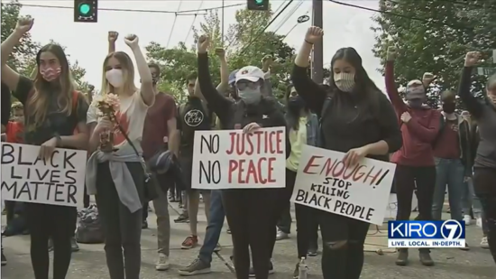 A KIRO 7 screenshot of a crowd of protestors standing a street with raised arms. They are holding painted signs that say Black Lives Matter and No Justice No Peace.