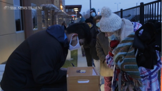 A video screenshot shows a line of masked people standing outside. They are approaching a masked person who is writing something on a table near a box of papers.