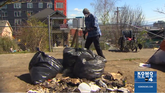 On a sunny day, black trash bags sit in a pile on the ground as a masked man stands nearby. A metal fence and a building are in the background.