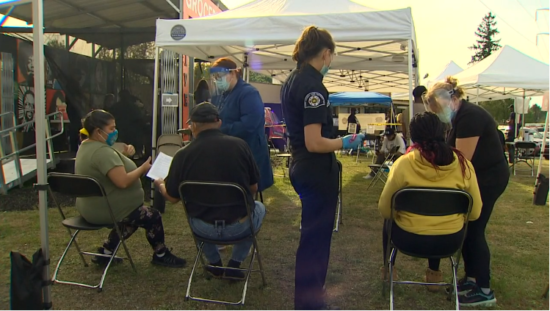 A row people sits in folding chairs on a lawn, with white canopies in the background and a dark blue uniformed medical worker standing between them.