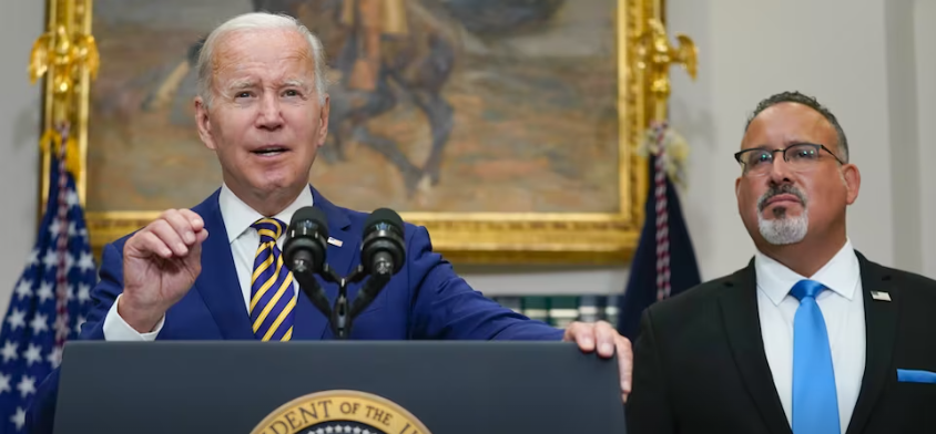 President Joe Biden stands at a podium next to U.S. Secretary of Education Miguel Cardona 