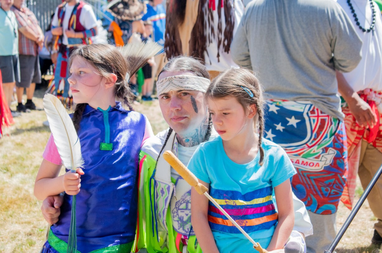 Ryan Wixon (center) looks at the camera as he takes a break from drumming. His daughters, Ahyoka, 10, (left) and Lily, 7, (right) gather around him. (Grace Deng/Washington State Standard)