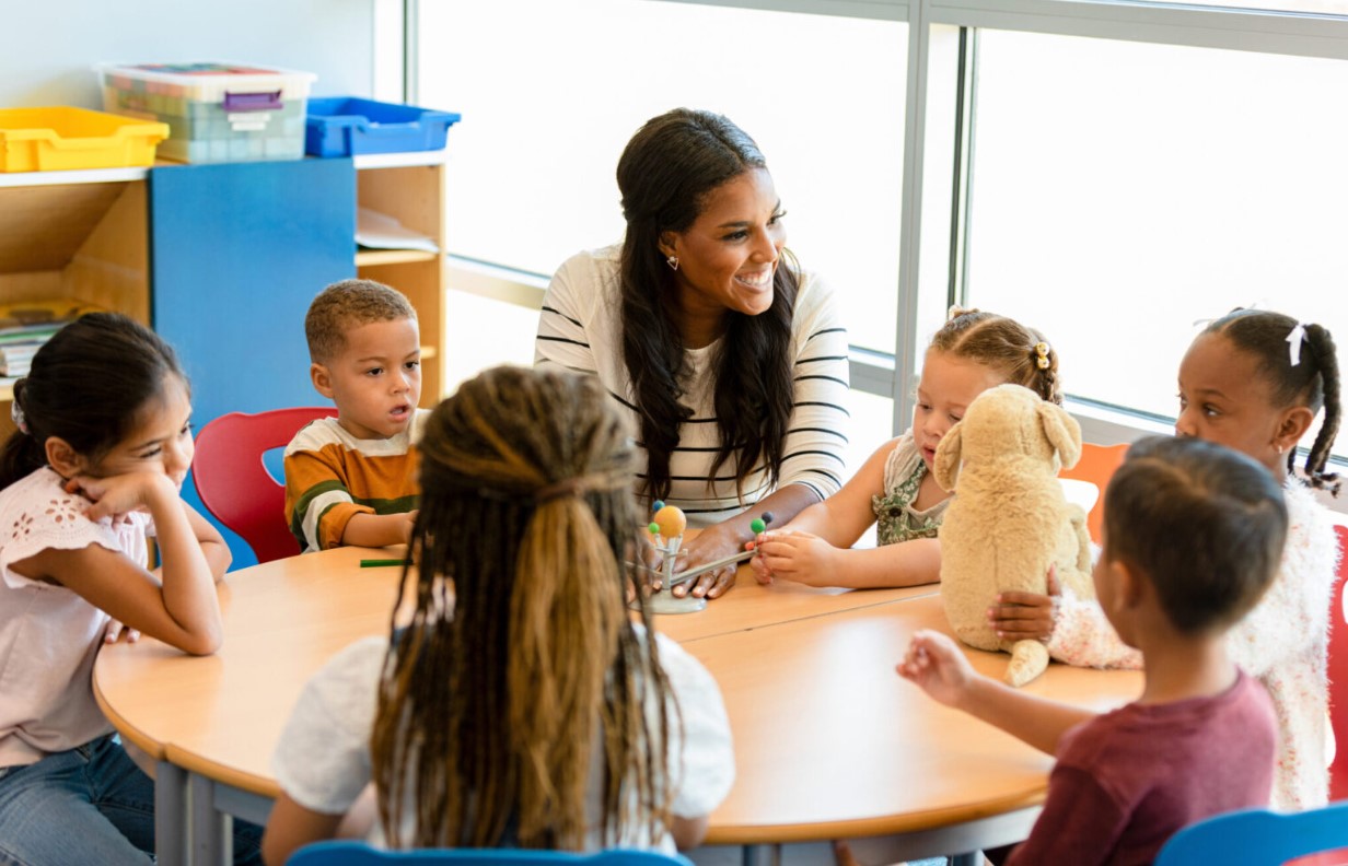 Teacher sits at a table with preschool students.