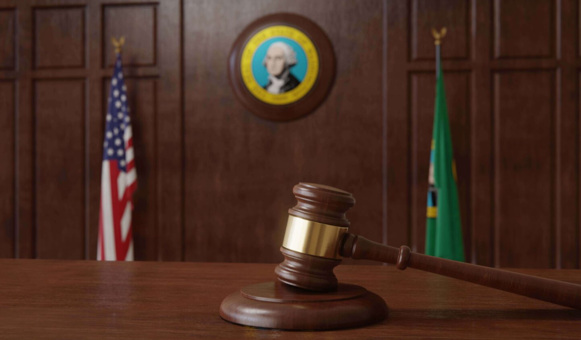 A judge’s gavel laying on a desk with a blurred image of the WA state seal and WA and American flags behind it.