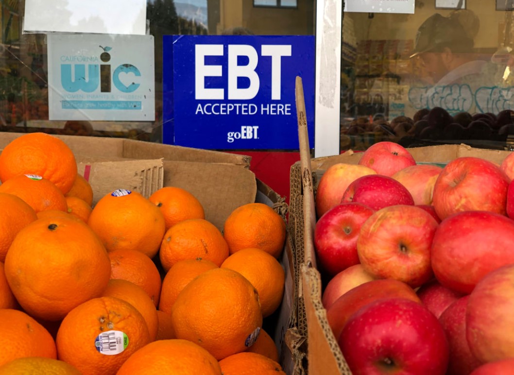 Photo of oranges and apples in boxes outside a storefront with a sign on the window that says “EBT accepted here”.