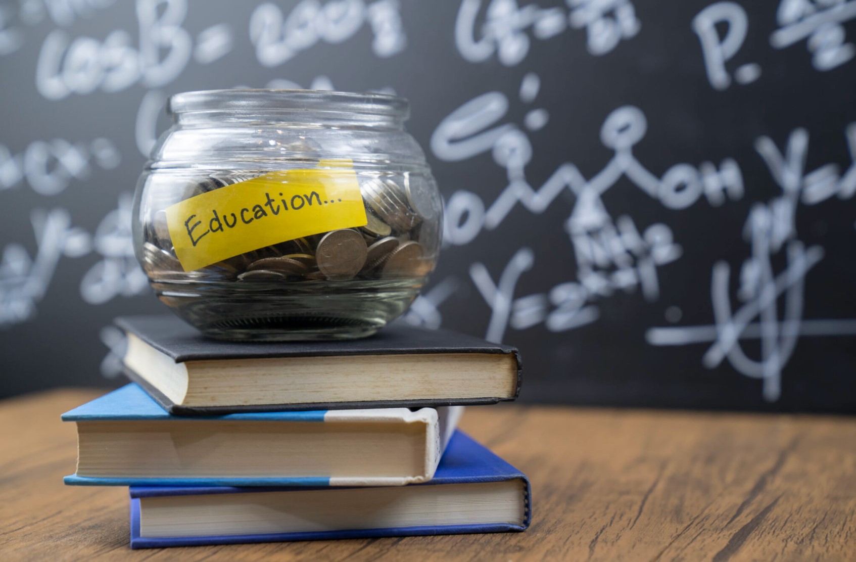 Stack of books in front of a chalkboard with a bowl on top filled with quarters and a yellow slip of paper with the word education written on it.