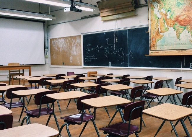 Empty classroom with wooden desktops, chalkboards on right side of room, white board at front of room.