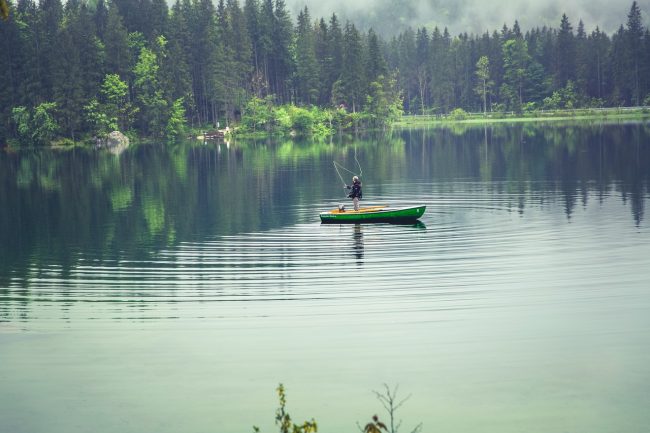 man standing up in small boat in middle of lake fishing; lake surrounded by evergreens