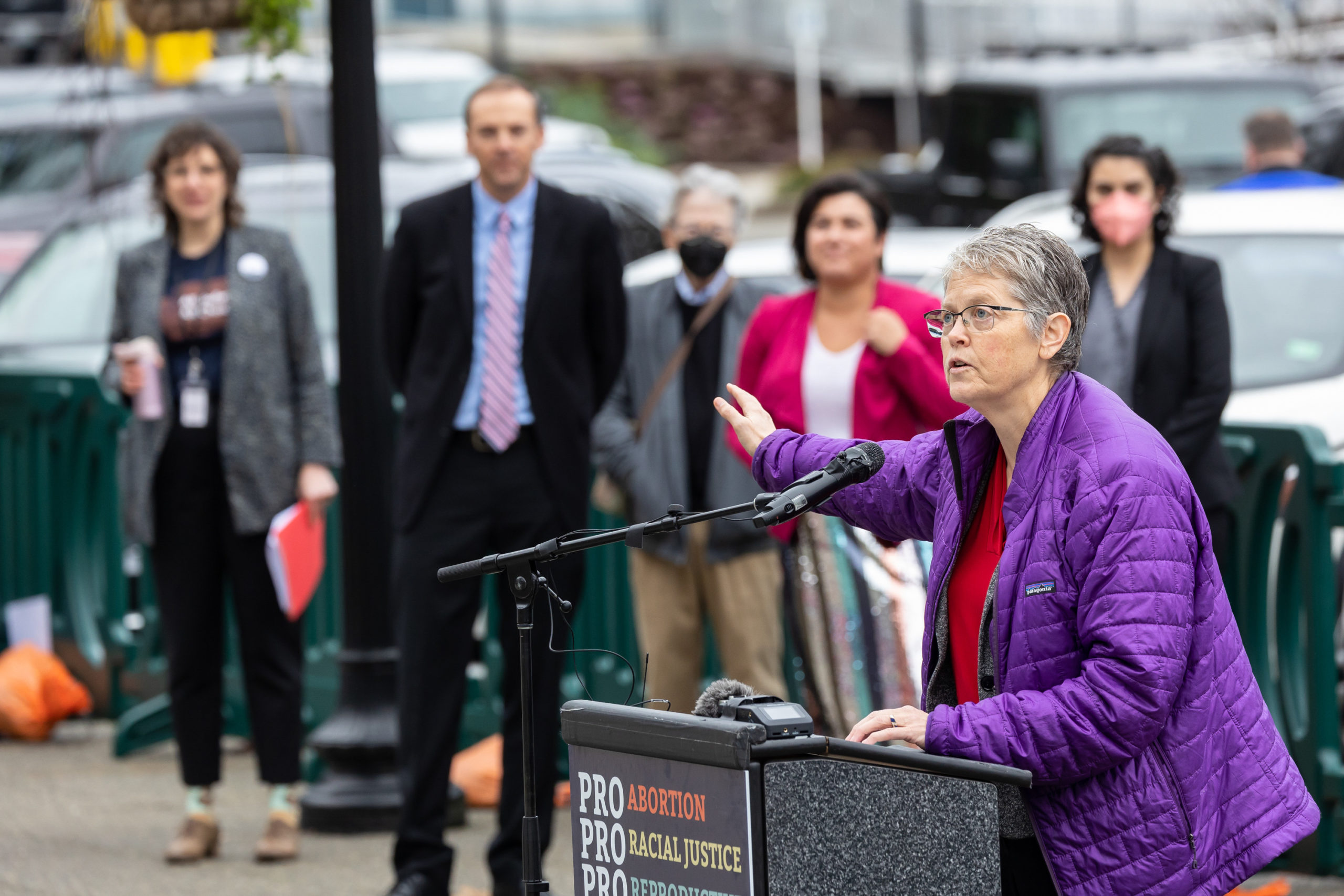 Speaker Jinkins in a purple puffy jacket standing in front of a podium, speaking and gesturing with her hands