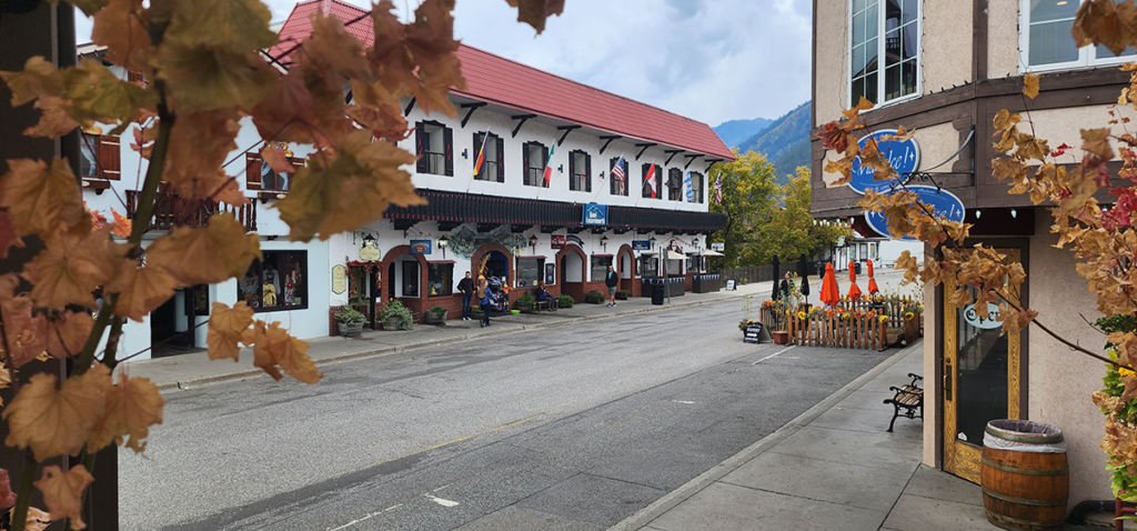 A scenic shot of a street in Leavenworth, Washington. In the foreground is a beige building with two blue signs and in the background a white building with a red roof and dark trim.