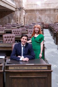 Rep. Alex Ramel sitting at his desk on the House floor, his Legislative Assistant Amanda is standing next to him.