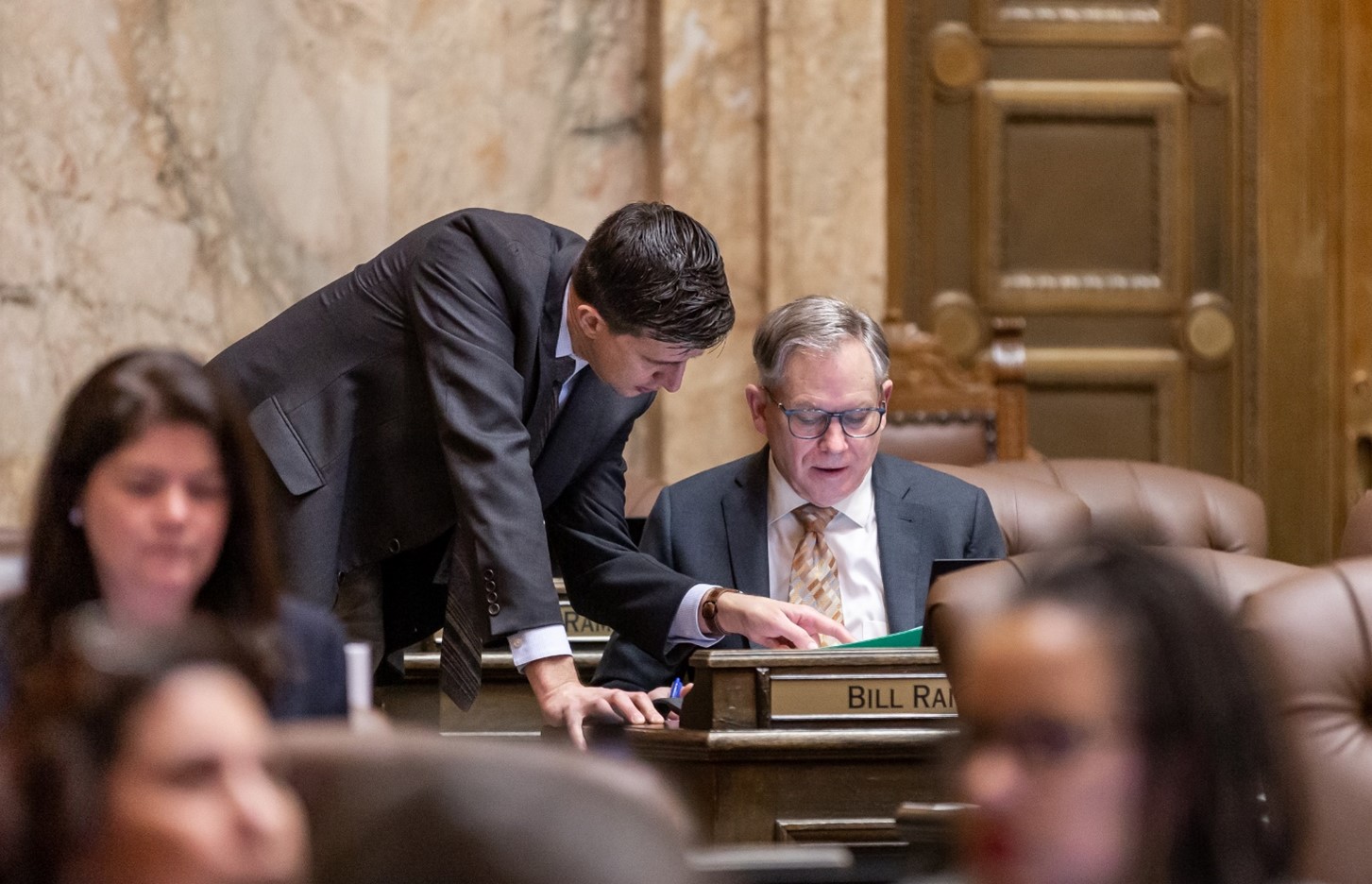 Rep. Ramel standing over a sitting Rep. Ramos at his desk on the House floor. Ramel stands to his right. Both are wearing gray suits, and are deeply focused on the topic at hand.