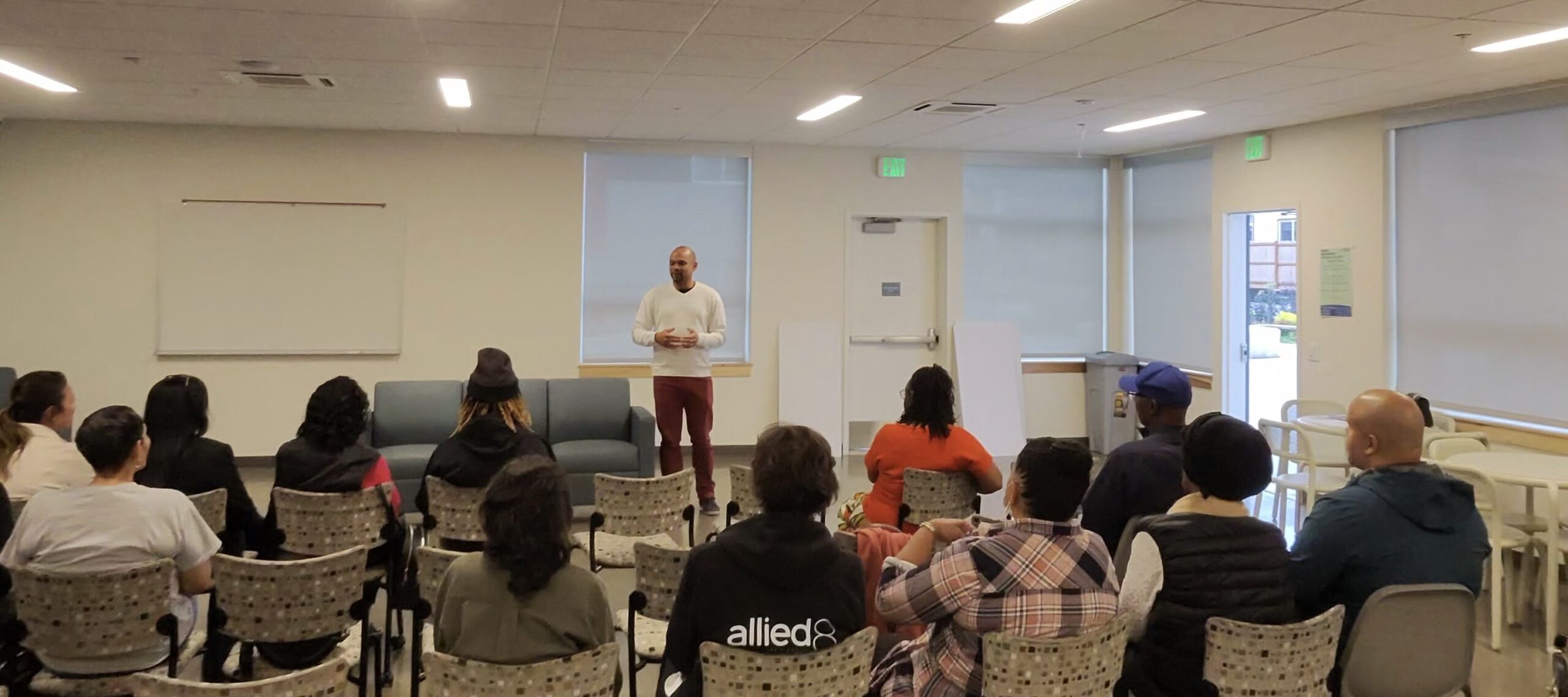 A man stands at the front of a room speaking to a small crowd of people who are seated in chairs.