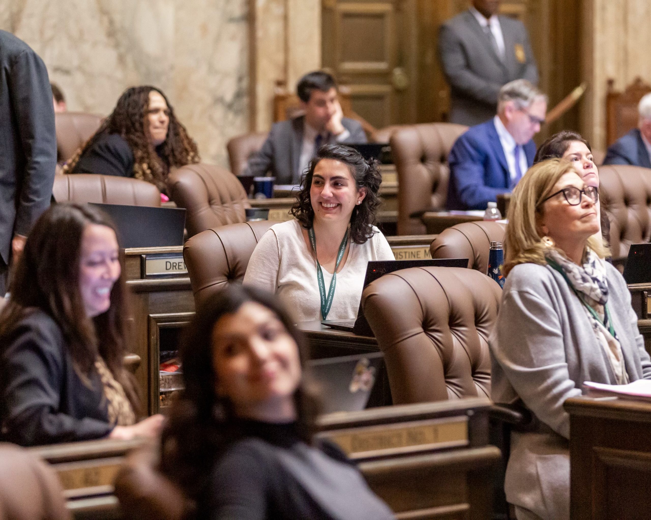 Rep. Farivar sitting at her desk among her colleagues on the House floor. She is looking to her right and smiling with her colleagues. The background of the image is blurred while she is in focus.